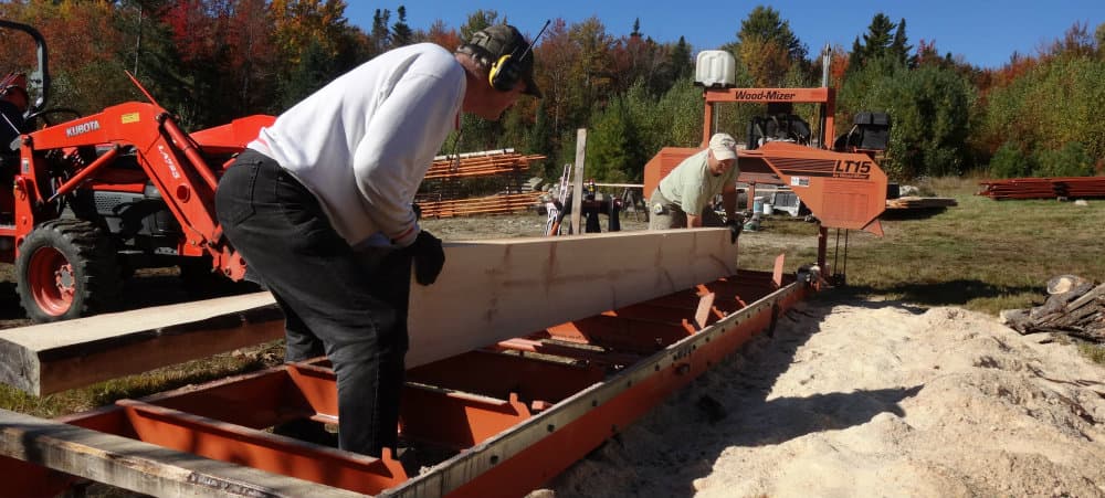 Two individuals operate a portable sawmill in a wooded area, processing a large wooden beam.