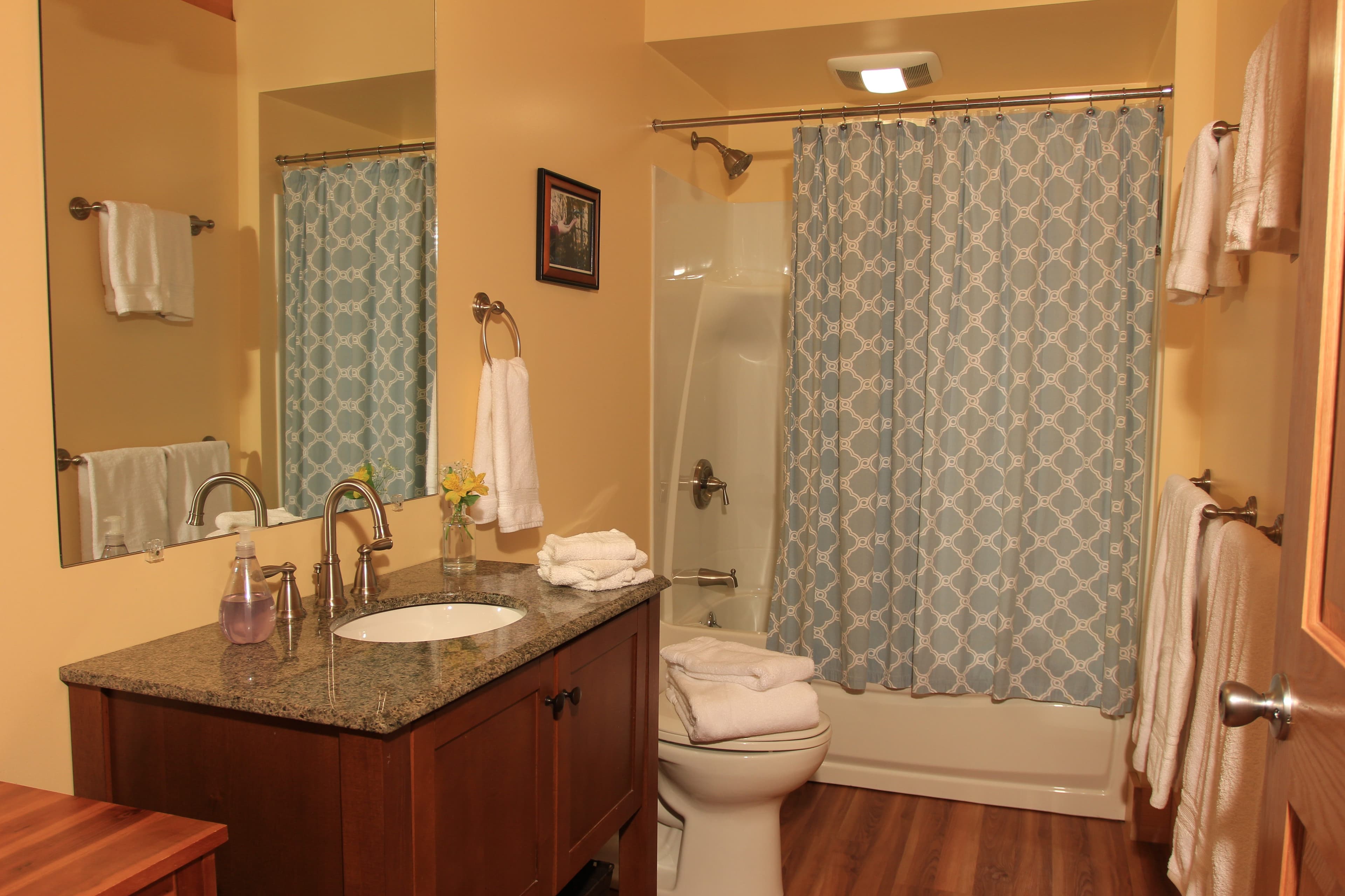 A modern bathroom features a dark wood vanity with a granite countertop, a large mirror, and a white sink. The space includes a toilet and a shower-tub combination enclosed by a blue and white patterned shower curtain, all set against warm tan walls and wood flooring.