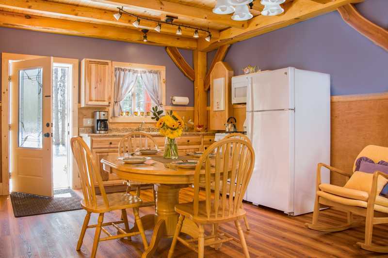 The interior of a rustic cabin features a kitchen and dining area with periwinkle blue walls, warm wood wainscoting, and exposed timber ceiling beams. A round wooden dining table with four matching chairs sits in the foreground, illuminated by a classic chandelier. The kitchen in the background includes light wood cabinetry, a white refrigerator, and a window with light-colored curtains, all set against wood flooring.