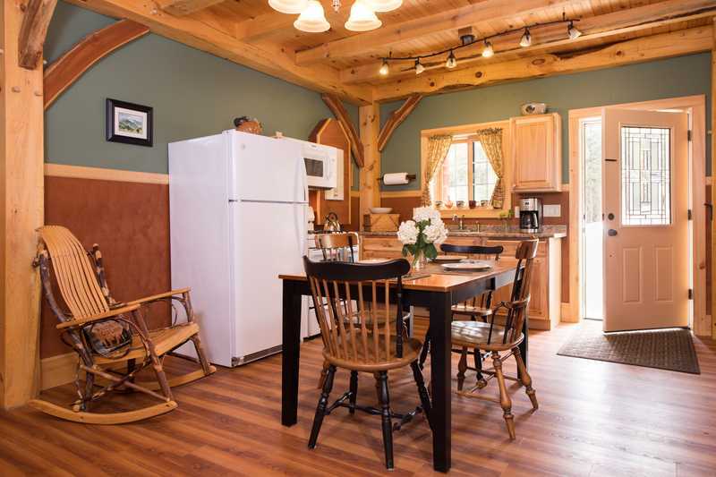 A cozy kitchen and dining area in a rustic cabin featuring light wood cabinetry, a white refrigerator, and a dark wooden dining table with black chairs. The room is detailed with exposed timber beams, warm wood-look flooring, and a wooden rocking chair in the corner, with a glass-paned door leading outside.