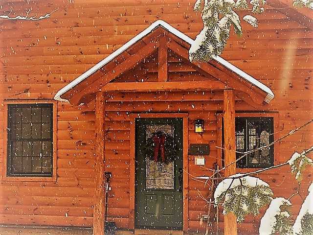 An up-close view of the entrance to a wooden timber-frame cabin at Robert Frost Mountain Cabins in Vermont, featuring a gabled porch roof and a festive holiday wreath on the front door, partially framed by snow-covered evergreen branches.