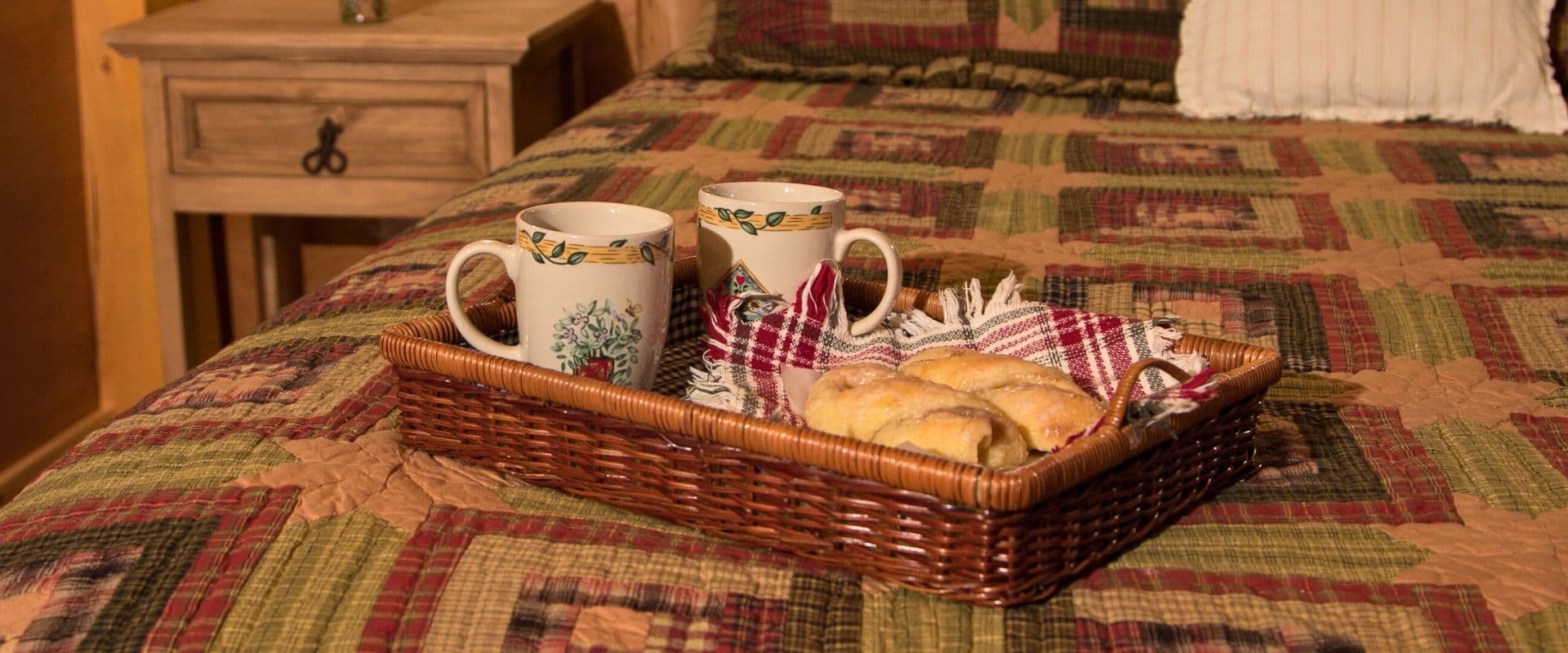 A woven tray with two cups and pastries sits on a bed with a patterned quilt.