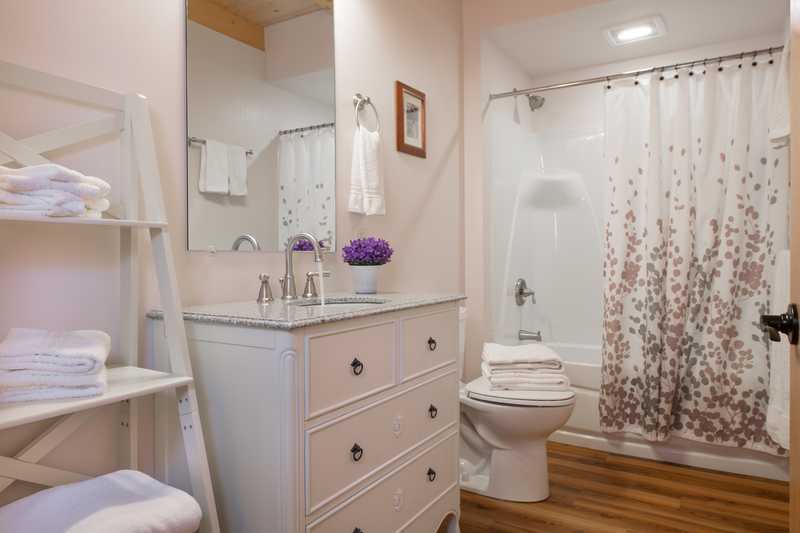 A bathroom inside a rustic cabin featuring a white dresser-style vanity with a silver faucet and a large wall mirror. The room has light pink walls, wood-look flooring, and a shower-tub combination with a white shower curtain featuring a delicate floral or branch pattern.