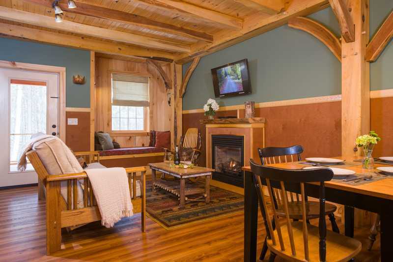 An open-concept living area inside a rustic cabin features warm wood flooring, exposed timber ceiling beams, and blue-gray walls with wood wainscoting. The space includes a wooden armchair with a white throw, a central coffee table on an area rug, a dining table with black chairs in the foreground, and a cozy window seat and fireplace with a mounted TV in the background.