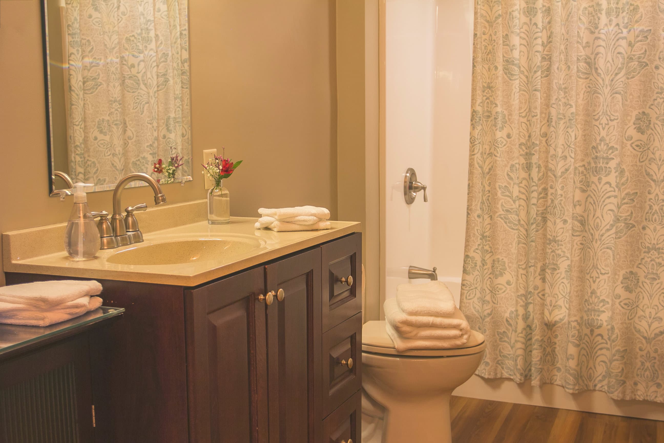A warm, inviting cabin bathroom featuring a dark wood vanity with a beige countertop and integrated sink. The space is accessorized with fresh white towels, a small vase of flowers, and a large mirror, while a patterned shower curtain adds a decorative touch to the room's calm, neutral tones.