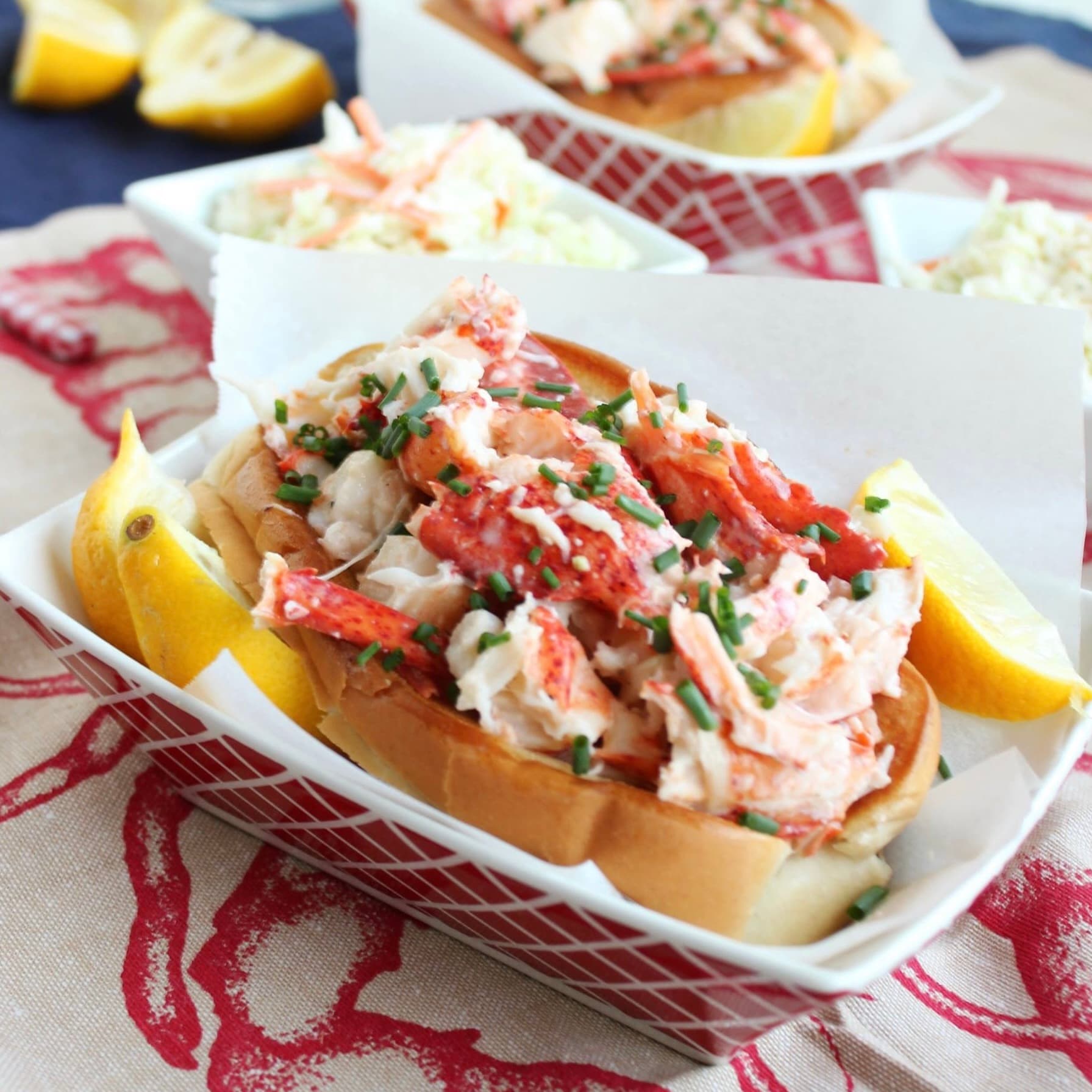 A lobster roll topped with chives and accompanied by lemon wedges, served in a red and white basket.