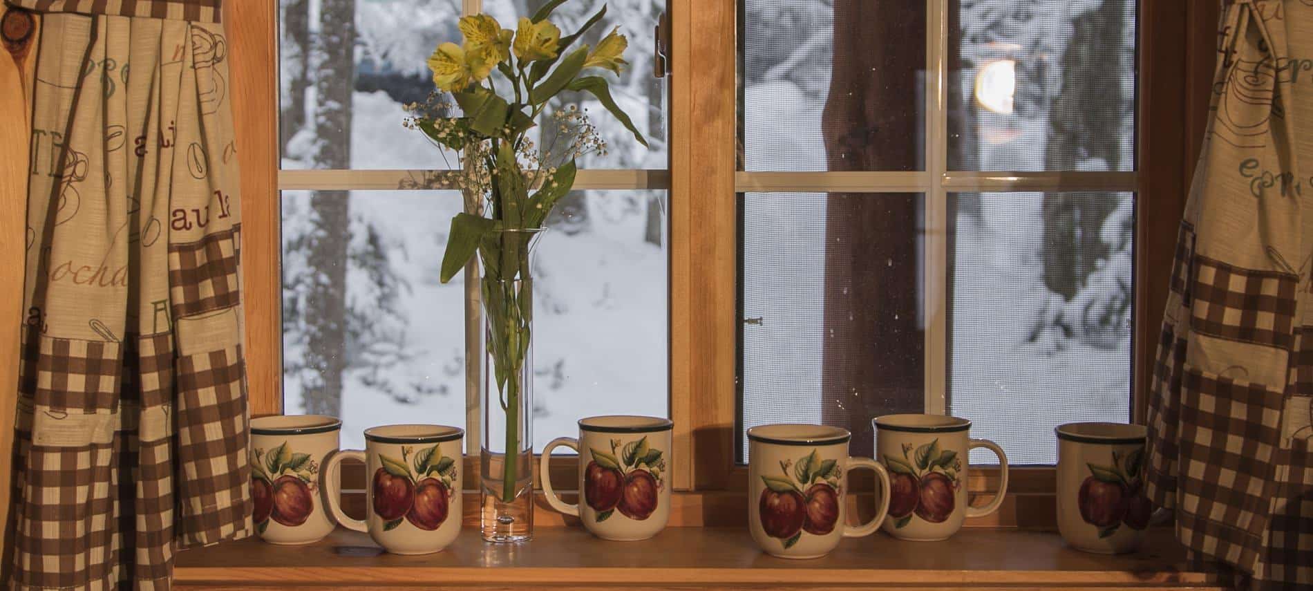 Window with checkered curtains, ceramic mugs with painted apples on the sill and a tall slender glass vase with yellow flowers