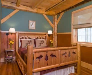 Cabin guest room, rustic wooden carved bed, two nightstands with lamps, blue-green walls, wood floors and window with shade