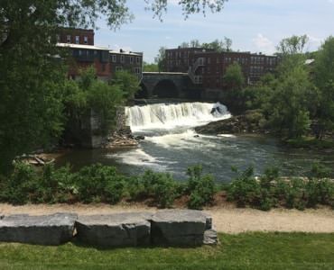 Short waterfall into a river surrounded by trees, plants, brick buildings and a bridge