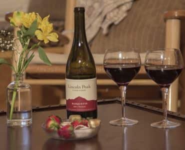 Bottle of wine, two wine glasses filled with red wine, glass jar with yellow flowers and bowl of chocolate covered strawberries