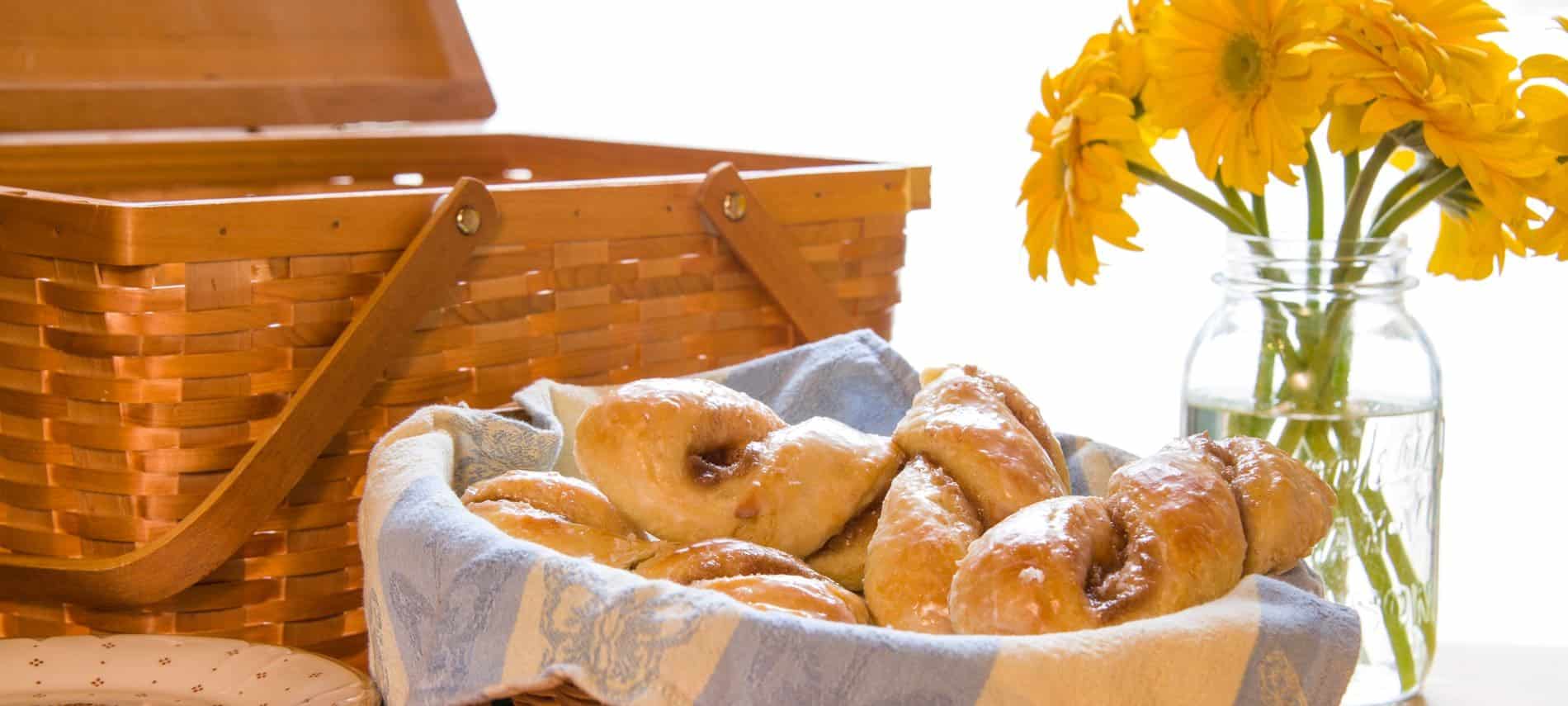 Wooden picnic basket next to a basket of fresh glazed twisted rolls and a glass Mason jar filled with bright yellow flowers