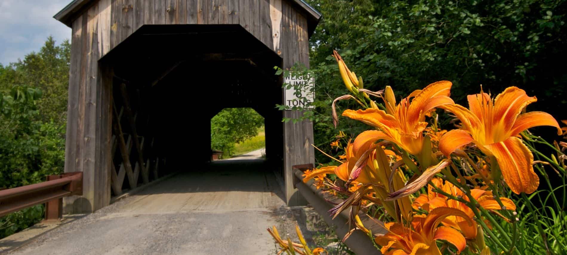 Wooden covered bridge surrounded by lush green trees and bright orange lilies in the foreground