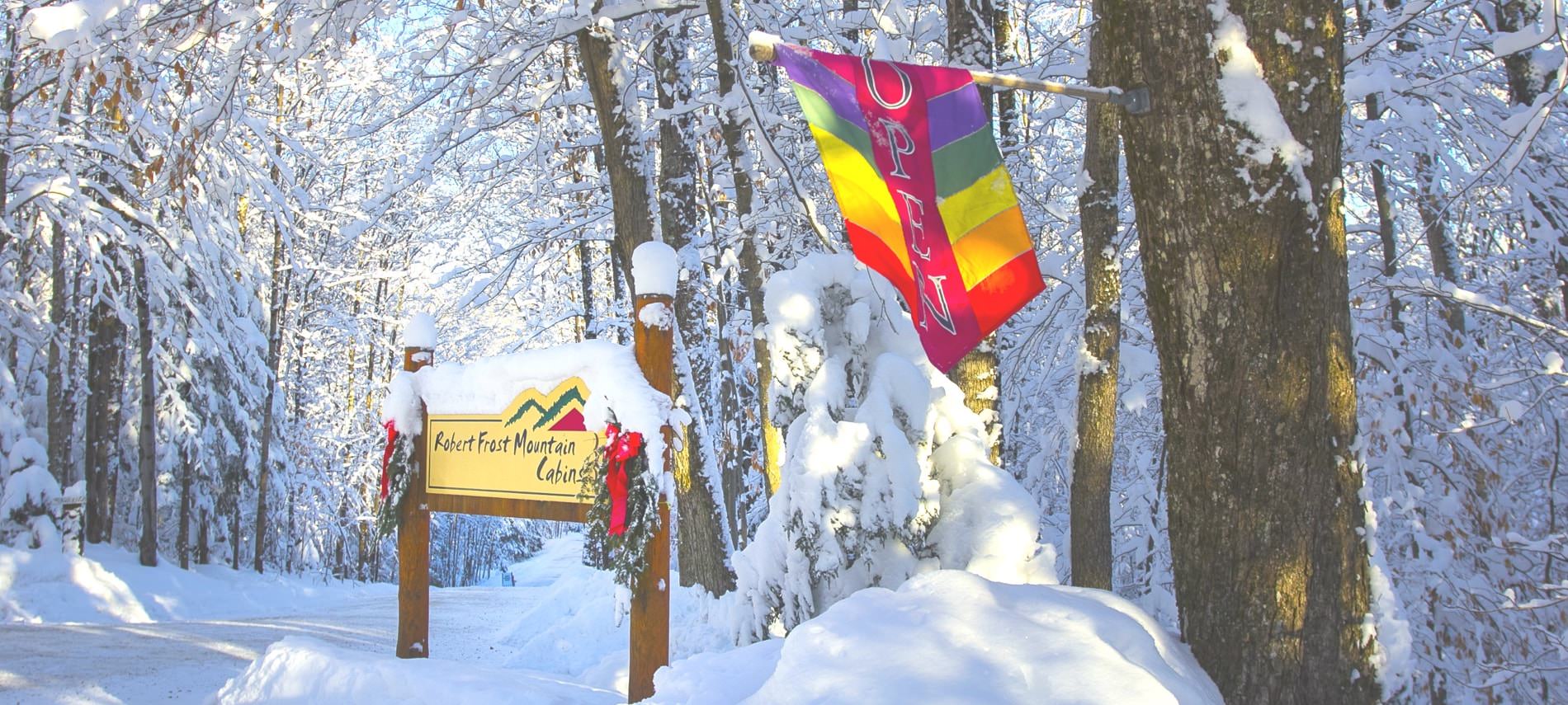 Entrance, wood and yellow sign that reads Robert Frost Mountain Cabins, colorful open flag, all surrounded by white snow covered trees and ground