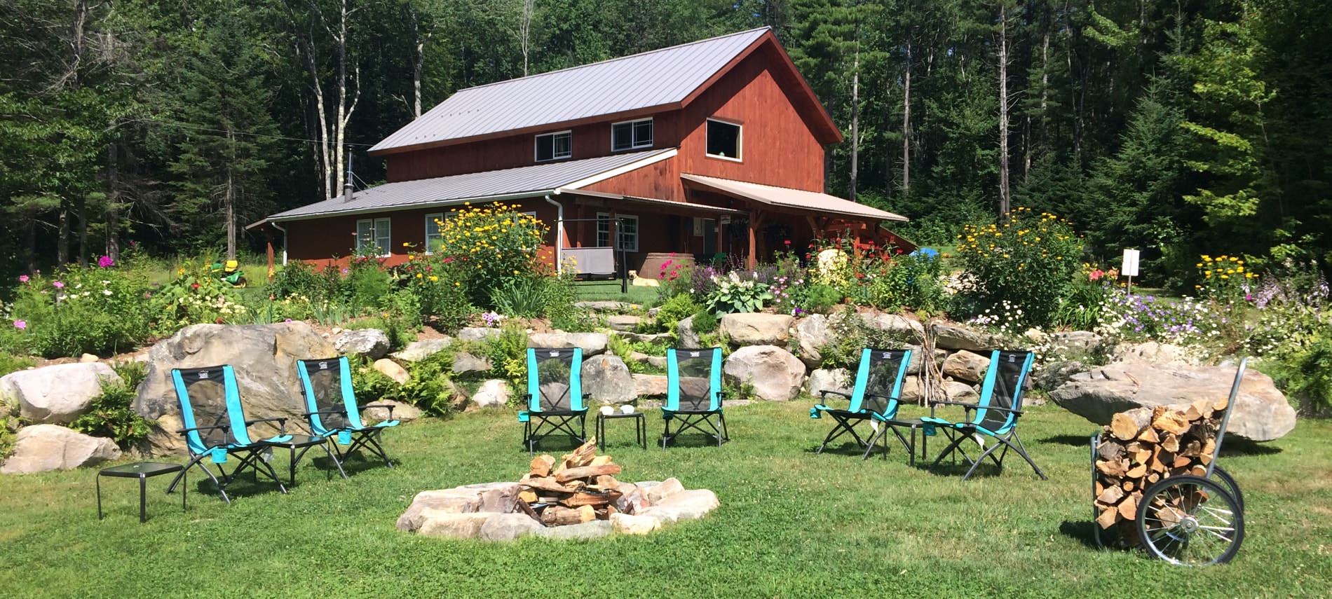 Camping chairs around a camp fire pit in the grass with flowers, boulders, tall trees and a large cabin in the background