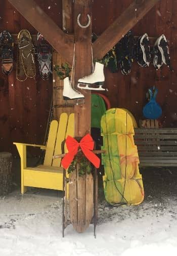 Side of a cabin with hanging pairs of snowshoes, propped up sleds, and a pair of hanging white ice skates