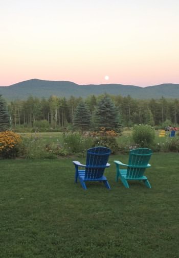 Two Adirondack chairs, one blue and one turquoise, on green grass overlooking pine trees, and the sun setting behind distant hills
