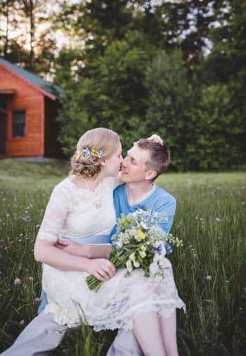 Woman sitting on a man's lap in the grass holding fresh flowers with green trees and a cabin in the background